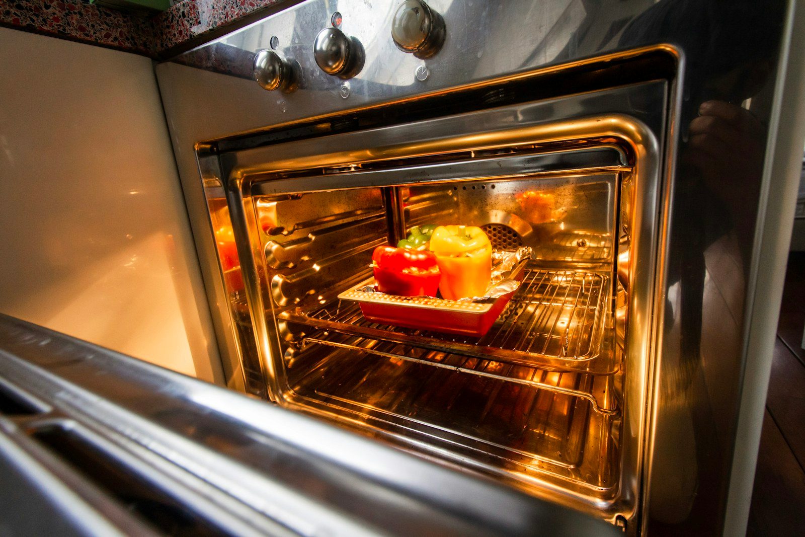 Open stainless steel oven with the light on and capsicums on a tray inside